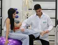 Estudante de Fisioterapia da Unoeste atende paciente sentada sobre bola de pilates roxa. Ambos interagem enquanto o aluno manipula a perna da paciente para exercício ou avaliação. Ao fundo, há halteres coloridos, esqueleto anatômico e ambiente de clínica bem equipado.