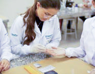 Aluna de Fisioterapia em jaleco branco e luvas coletando amostra de sangue do dedo de um colega, utilizando lancetas e kits de teste em bancada de laboratório.