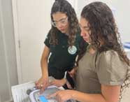 Duas estudantes, uma com camisa polo verde escuro da Unoeste e outra com blusa bege, analisam juntas um equipamento médico sobre uma bancada. Uma delas aponta para o visor do aparelho enquanto a outra segura um celular, ambas com cabelos cacheados e longos.