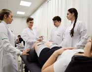 Alunos do curso de Fisioterapia da Unoeste, em jalecos brancos, realizam procedimento com eletrodos aplicados nas coxas de uma paciente deitada. Uma estudante opera o aparelho enquanto os demais observam atentos. A sala é clara, com cortinas brancas e teto com iluminação forte.