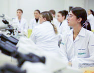 Aluna de Fisioterapia em jaleco branco e óculos prestando atenção em aula prática de laboratório, com microscópios em primeiro plano e outros alunos ao fundo.