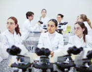 Grupo de alunos de Fisioterapia em jalecos brancos assistindo a uma aula ou apresentação em laboratório, com microscópios nas bancadas e focados na instrução.