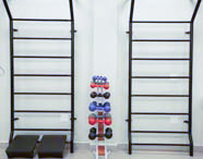 Ambiente de Fisioterapia com espaldar para exercícios, suporte com pesos coloridos (halteres e kettlebells) e bancada com pia, indicando um espaço de reabilitação.