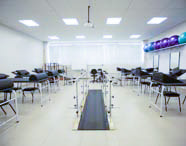 Ampla sala de fisioterapia ou laboratório de reabilitação com passarela de marcha com barras paralelas, várias macas de tratamento, bolas suíças e espelhos, pronta para atividades práticas.