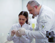 Professor de Fisioterapia em jaleco branco orientando uma aluna em luvas, mostrando uma pequena amostra ou material para análise em atividade prática.