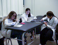 Três alunas de Fisioterapia em jalecos brancos estudando e fazendo anotações em tablets, sentadas em uma mesa de laboratório com foco em suas atividades.