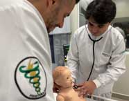 Estudante de Fisioterapia da Unoeste, com jaleco branco, utiliza estetoscópio para auscultar manequim infantil em laboratório de simulação. Outro aluno observa de perto. Ao fundo, painel eletrônico simula sinais vitais, destacando a tecnologia empregada no ensino prático.