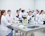 Alunos de Fisioterapia em jalecos brancos sentados em mesas de laboratório equipadas com microscópios, participando de uma aula prática ou teórica com atenção.