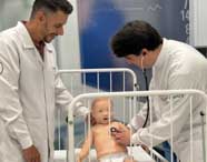 Em laboratório de simulação da Unoeste, professor e estudante de Fisioterapia, ambos de jaleco branco, realizam exame com estetoscópio em manequim infantil dentro de berço hospitalar. O cenário reproduz ambiente pediátrico, com paredes azuis e detalhes hospitalares.
