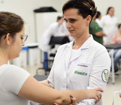 aluna do curso de Fisioterapia da Unoeste, identificada pelo jaleco branco com nome bordado, durante atividade prática em laboratório. Segurando no braço para demonstrar técnica de avaliação ou reabilitação motora, em ambiente acadêmico com outros alunos ao fundo.