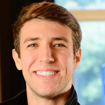 Vitor, egresso de Educação Física - Bacharelado, posando em um ambiente interno com uma janela ao fundo. Ele tem cabelos curtos e um sorriso largo.