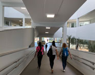 Três estudantes universitárias, duas de cabelo longo e uma de cabelo cacheado, caminham por uma rampa interna acessível, segurando máscaras e mochilas. O espaço é amplo, iluminado, com corrimãos metálicos e paredes brancas. Plantas decoram o interior do prédio moderno da Unoeste, campus Jaú, voltado ao curso de Educação Física.