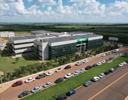 Vista aérea do campus da Unoeste em Jaú, mostrando edifício moderno com fachada de vidro, painéis solares no telhado e jardins bem cuidados. Diversos carros estacionados ao redor. Ao fundo, paisagem rural com plantações e céu parcialmente nublado. Estrutura destaca a infraestrutura do curso de Educação Física.