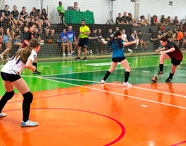 Quadra poliesportiva coberta durante competição feminina de futsal entre estudantes do curso de Educação Física da Unoeste Jaú. Atletas com uniformes esportivos jogam enquanto público assiste nas arquibancadas. Piso da quadra é verde e laranja, redes de proteção ao fundo e árbitro acompanha o jogo.