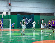 Partida masculina de futsal entre estudantes do curso de Educação Física da Unoeste Jaú. Jogadores com uniformes verdes, brancos e roxos se posicionam próximos ao gol, enquanto torcida acompanha nas arquibancadas. Rede de proteção cobre o fundo, quadra verde e laranja, banner decorativo pendurado na parede.