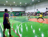 Jogo de futsal masculino em andamento no ginásio da Unoeste Jaú. Jogador de uniforme amarelo prepara-se para entrar, enquanto goleiro defende chute à gol. Outros atletas em quadra, público atento nas arquibancadas e árbitros observam. Piso verde e laranja, rede de proteção e banner institucional ao fundo.