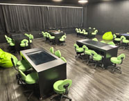 Sala de tecnologia educacional da Unoeste Jaú, com várias mesas digitais retangulares e cadeiras verdes ergonômicas. Puffs verdes espalhados pelo chão de madeira escura, cortinas pretas cobrindo as paredes. Espaço moderno e inovador, destinado a atividades interativas dos estudantes do curso de Educação Física.