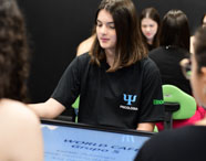 Aluna do curso de Psicologia da Unoeste Jaú participa de atividade interativa em sala moderna, vestindo camiseta preta com símbolo da Psicologia. Sentada à mesa digital sensível ao toque, junto a outros estudantes, utiliza tecnologia educacional para dinâmicas de grupo, promovendo aprendizado inovador e colaborativo.