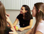 Três alunas de Psicologia em uma sala com decoração de casa simulada, interagindo e conversando em um ambiente descontraído, com frutas em uma fruteira na mesa.