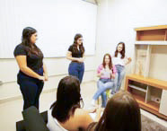 Grupo de alunas de Psicologia participando de uma atividade prática em uma sala que simula um ambiente residencial, com algumas em pé e outras sentadas, interagindo e observando.