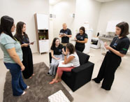 Sala simulada da Unoeste Jaú, onde estudantes de Psicologia, vestidos com camisetas do curso, observam e anotam durante dinâmica prática. Uma dupla simula atendimento sentada em sofá preto, enquanto colegas e professora acompanham. Ambiente realista, com móveis domésticos, promovendo aprendizagem ativa e humanizada.