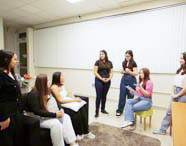 Alunas de Psicologia em uma aula prática ou simulação em um ambiente que se assemelha a uma sala de estar, com sofá, estante e persianas, todas engajadas na atividade.