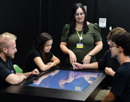 Grupo de estudantes do curso de Psicologia da Unoeste Jaú reunidos em torno de mesa digital interativa, acompanhados por professora com crachá. Todos atentos à tela sensível ao toque, participando de dinâmica tecnológica em sala escura, cadeiras verdes ao fundo. Atividade estimula uso de recursos digitais no ensino superior.