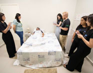 Estudantes de Psicologia da Unoeste Jaú participam de simulação em ambiente que reproduz quarto residencial. Um aluno deitado na cama, outros observam e fazem anotações, acompanhados por professora. Todos usam camisetas do curso. Espaço decorado com quadros, móveis claros e iluminação suave, favorecendo o aprendizado prático.