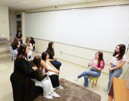 Alunas de Psicologia em um ambiente de casa simulada, interagindo e estudando em grupo, com uma estante e um refrigerador ao fundo, criando um cenário de prática realística.