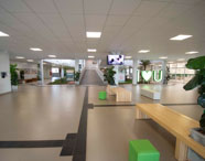 Hall de entrada amplo do campus Unoeste Jaú, com bancos de madeira, pufes verdes, plantas ornamentais e sinalização tátil no piso. Ao fundo, rampa de acesso, jardim interno e escultura “I ♥ U”. Placas informativas e TV suspensa complementam o espaço, projetado para conforto, acessibilidade e convivência dos estudantes.