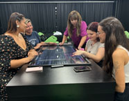 Estudantes do curso de Psicologia da Unoeste Jaú utilizam mesa digital interativa em laboratório tecnológico. Em volta da tela sensível ao toque, grupo discute conteúdos exibidos, como imagens e gráficos. Sala moderna, cadeiras verdes e cortinas pretas ao fundo, mostrando o uso de tecnologia avançada no ensino.
