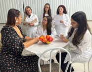 Estudantes de Psicologia da Unoeste Jaú em aula prática, usando jalecos brancos e cadernos, simulam atendimento em ambiente de consultório. Sentados à mesa com fruteira de frutas artificiais, observam e registram informações, integrando teoria e prática em cenário realista de formação profissional.