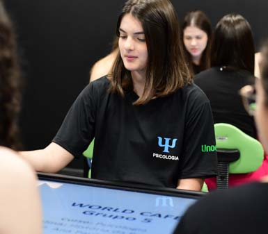 Estudante do curso de Psicologia da Unoeste Jaú sentada em frente a uma mesa digital interativa, usando camiseta preta com símbolo da Psicologia e logotipo da instituição na manga. Ao redor, outros alunos participam da atividade em ambiente moderno, com cadeiras verdes e parede escura, demonstrando integração e tecnologia no ensino.