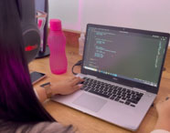 Aluna do curso de Ciência de Dados utiliza notebook sobre mesa de madeira, programando com fones de ouvido grandes. Ao lado, garrafa rosa, celular e objetos pessoais. O ambiente é iluminado e organizado, refletindo o estudo individual em tecnologia na Unoeste.