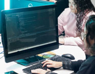 Duas alunas da Faculdade de Ciência de Dados trabalham em computadores de mesa, concentradas em códigos exibidos nas telas. Uma delas usa óculos e blusa escura, a outra veste moletom claro. O laboratório possui iluminação moderna e monitores grandes, promovendo aprendizado prático.