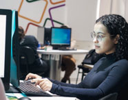 Aluna do curso de Ciência de Dados digita no teclado de computador em laboratório de informática da Unoeste. Ela usa blusa de manga longa azul-marinho e óculos, sentada à mesa branca em ambiente iluminado, com outros estudantes ao fundo também envolvidos em atividades digitais