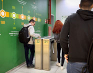 Estudantes entram pela catraca numerada em corredor interno da faculdade, ao lado de parede verde com linha do tempo dos cursos de tecnologia da Unoeste. Jovens usam mochilas e roupas casuais, movimentando-se para acessar as dependências do prédio de Ciência de Dados