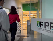 Dois estudantes caminham pelo corredor de entrada da FIPP – Faculdade de Informática de Presidente Prudente, identificada por adesivo na porta de vidro. Ao fundo, vitrine iluminada exibe equipamentos antigos de computação, compondo o cenário institucional e tecnológico.