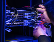 Pessoa manuseia fios e conexões em rack de servidores iluminado por luz azul, ajustando cabos pretos e vermelhos. A cena mostra parte da infraestrutura de TI utilizada nos cursos de Ciência de Dados da Unoeste, ressaltando a prática em ambientes reais de tecnologia.
