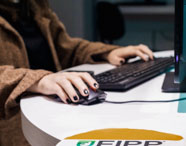 Estudante da Faculdade de Ciência de Dados opera mouse e teclado em mesa redonda, com monitor à frente. Veste casaco marrom e esmalte preto, enquanto realiza tarefa prática. 