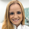 Gabrielle, aluna do curso de biomedicina no campus Guarujá, trajando jaleco branco. O fundo apresenta armários brancos e equipamentos médicos, sugerindo um laboratório acadêmico bem organizado. A iluminação suave cria um ambiente acolhedor e propício ao aprendizado.
