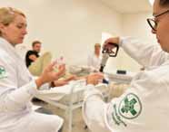 Curso de Biomedicina:  Professora loira de jaleco branco, com bordado verde, orienta aluna que utiliza equipamento de ventosaterapia. Ambiente de sala prática, paredes brancas, leitos ao fundo e outros estudantes participando da atividade, reforçando o contexto de ensino em saúde e bem-est