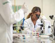 Curso de Biomedicin:  Aluna de cabelos castanhos claros, presa em rabo, com jaleco branco Unoeste, trabalha em laboratório moderno, manuseando microscópio óptico. Tubos de ensaio coloridos e equipamentos de vidro estão sobre a bancada, compondo um cenário de aula prática, limpo e organizado.