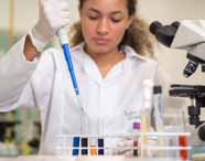 Faculdade de Biomedicina. Jovem estudante com cabelos cacheados presos e jaleco branco manipula micropipeta azul para transferir líquido para tubos de ensaio transparentes, posicionados em suporte branco ao lado de um microscópio óptico. O ambiente é um laboratório com bancada cinza e equipamentos científicos ao fundo.