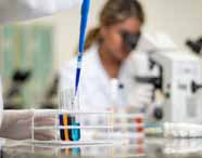 Faculdade de Biomedicina. Mão de estudante de jaleco branco utiliza micropipeta azul para adicionar líquido azul em tubos de ensaio, próximos a tubos com líquidos laranja e amarelo. Ao fundo, aluna com cabelos presos trabalha em bancada de laboratório equipada com microscópios, evidenciando