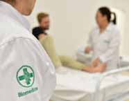 Faculdade de Biomedicina. Em sala branca de simulação, três pessoas com jaleco branco atendem paciente masculino loiro sentado em maca hospitalar. O foco está no detalhe do jaleco, com símbolo verde do curso de Biomedicina no braço, reforçando o ambiente acadêmico e prático.