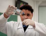 Aluno de Biomedicina em jaleco branco e luvas despejando líquido de uma garrafa em uma proveta em um laboratório, realizando um experimento.