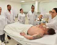 Grupo de cinco alunos em jaleco branco observam procedimento de ventosaterapia. Paciente deitado, tatuagem no braço, sobre maca coberta por lençol branco. Ambiente claro com paredes bege e móveis brancos. Equipamento de ventosa transparente aplicado nas costas do paciente.