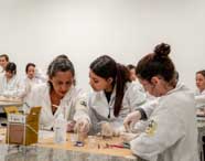 Três alunas de Biomedicina em jalecos brancos e luvas, realizando atividades práticas com vidraria de laboratório e materiais de pesquisa em uma bancada.