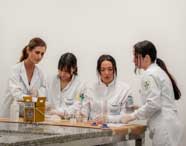 Quatro alunas de Biomedicina em jalecos brancos e luvas, concentradas em uma atividade prática em laboratório, com vidrarias e equipamentos científicos na bancada.